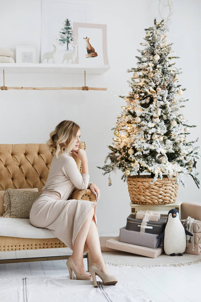 Beautiful platinum blonde model girl with perfect body in knitted dress posing near Christmas tree in interior. Young slim model woman in trendy outfit sits on a couch in New year interior. - 写真・画像