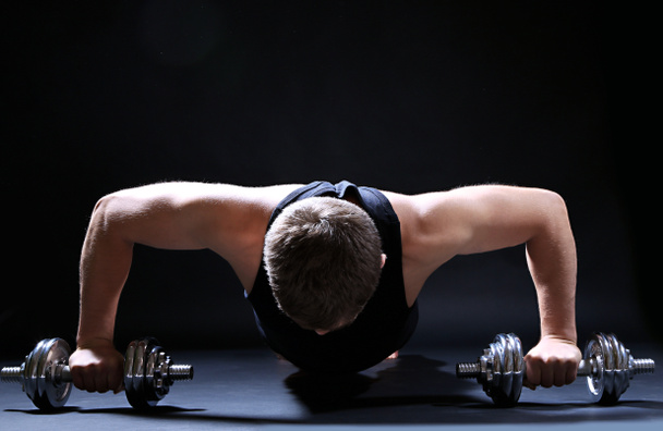 Красивый молодой мускулистый спортсмен выполняет упражнения с гантелями на тёмном фоне
 - Фото, изображение