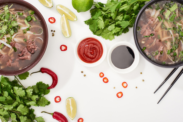 bovenaanzicht van pho in kommen bij eetstokjes, limoenen, chili en sojasauzen en koriander op witte achtergrond - Foto, afbeelding