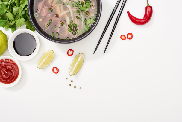 верхний вид фо в миске рядом палочки для еды, лайм, чили и соевые соусы и кориандр на белом фоне
 - Фото, изображение