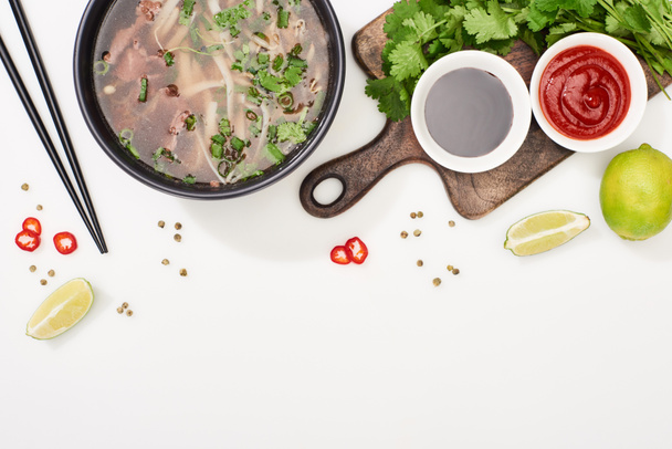 верхний вид фо в миске рядом палочки для еды, лайм, чили и соевые соусы и кориандр на белом фоне
 - Фото, изображение