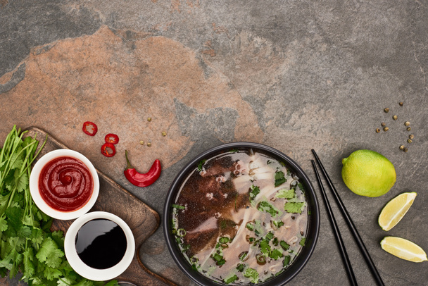 верхний вид фо в миске рядом палочки для еды, лайм, чили и соевые соусы и кориандр на каменном фоне
 - Фото, изображение