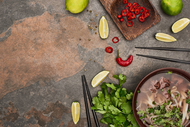 bovenaanzicht van pho in kom bij eetstokjes, limoen, chili en koriander op stenen ondergrond - Foto, afbeelding