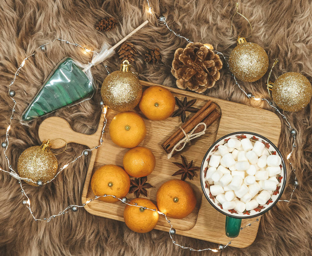 Wooden board with tangerines and cocoa with marshmallows, golden balls, cones, cinnamon and anice star, caramel on a background of brown fur. New Year flat lay. - Φωτογραφία, εικόνα