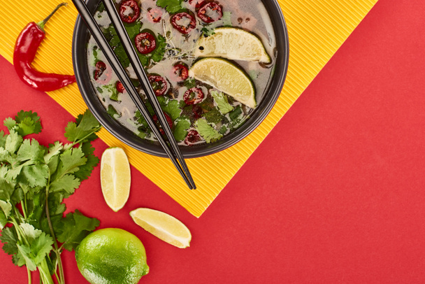 top view of pho in bowl with chopsticks near lime, chili and coriander on red and yellow background - Fotoğraf, Görsel