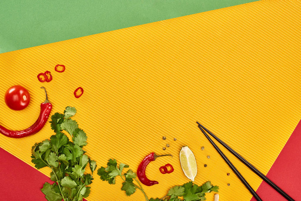 bovenaanzicht van eetstokjes, limoen, chili en koriander op rode, groene en gele achtergrond - Foto, afbeelding