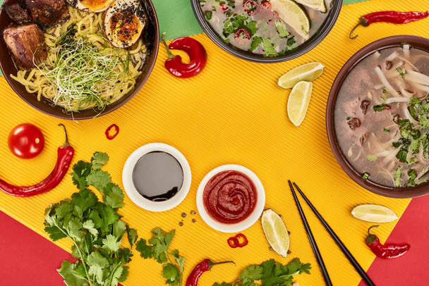 vrchní pohled na pho a ramen v miskách v blízkosti vápna, chilli a sójové omáčky a koriandru na barevném pozadí - Fotografie, Obrázek