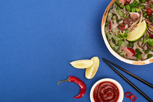 вид сверху на фо в миске рядом палочки для еды, лайм, соус чили на голубом фоне
 - Фото, изображение