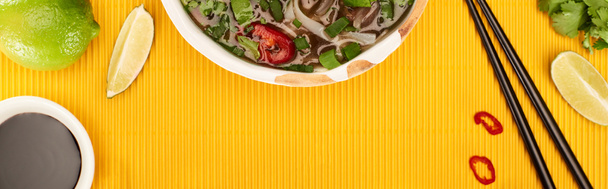 top view of pho in bowl near chopsticks, lime, soy sauce and coriander on yellow textured background, panoramic shot - Valokuva, kuva