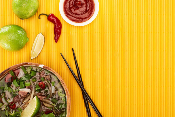 top view of pho in bowl near chopsticks, lime, chili sauce on yellow textured background - Фото, зображення