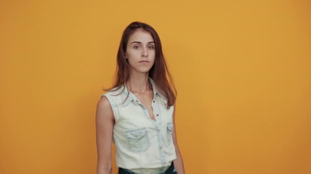 Strict caucasian young woman in blue denim shirt staying aside on orange wall - Imágenes, Vídeo