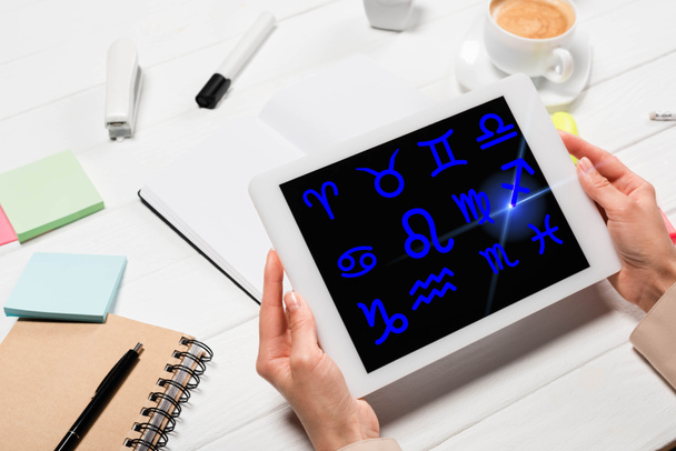 vista recortada de la mujer sosteniendo tableta digital con signos del zodiaco en el lugar de trabajo con suministros de oficina y café
 - Foto, Imagen