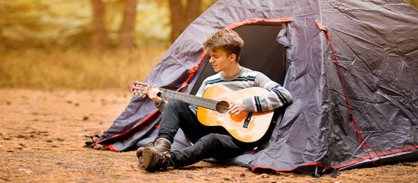 Banner θέα χαμογελαστός νεαρός άνδρας κάθεται κοντά τουριστική σκηνή και παίζει κιθάρα στο δάσος - Φωτογραφία, εικόνα