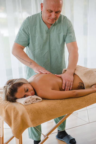 Mulher loira relaxante recebendo massagem nas costas do fisioterapeuta masculino no salão de beleza deitado na mesa de massagem. Jovem com tatuagens relaxantes no conceito de centro de spa - Close-up
 - Foto, Imagem