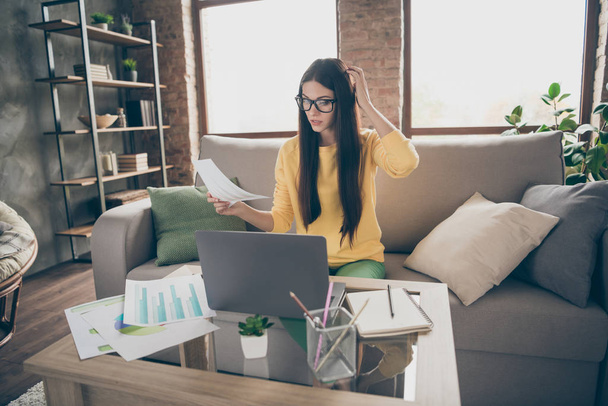 Doubtful uncertain girl worker marketer hold paper document decide decision try find startup solution sit divan wear yellow jumper in house indoors - Fotoğraf, Görsel