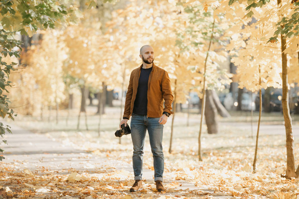 Photographe brutal chauve avec une barbe dans une veste en cuir de daim, chemise bleue, jeans et bottes Chelsea cherche quelqu'un dans le parc dans l'après-midi
 - Photo, image