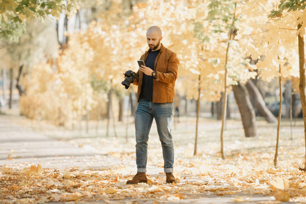 Bald stylish photographer with a beard in a suede leather jacket, blue shirt, jeans, and Chelsea boots holds the camera and looks for something in his smartphone in the park in the afternoon - Photo, Image