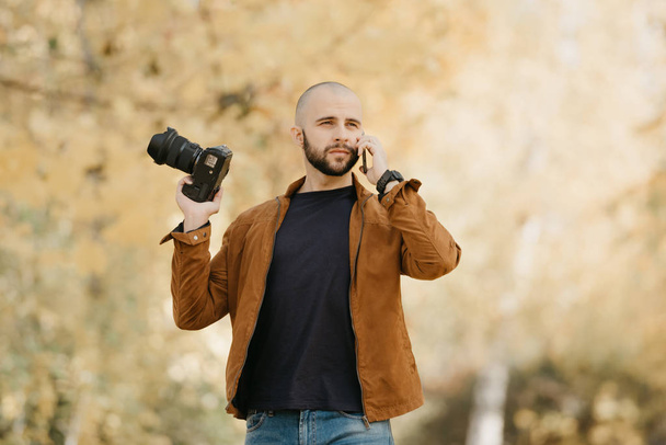Fotógrafo elegante careca com uma barba em uma jaqueta de couro de camurça, camisa azul, jeans com relógio de pulso segura a câmera na mão dobrada e chama alguém por seu smartphone no parque à tarde
 - Foto, Imagem
