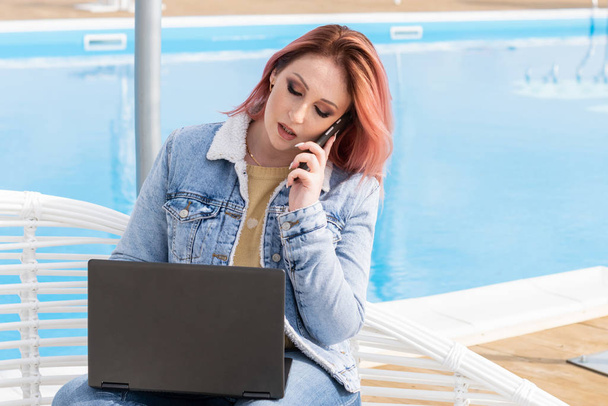 Kaunis tyttö farkut takki kannettavan tietokoneen lähellä uima-allas. tyylikäs freelancer meren rannalla. Punatukkainen tyttö. liikenainen
 - Valokuva, kuva