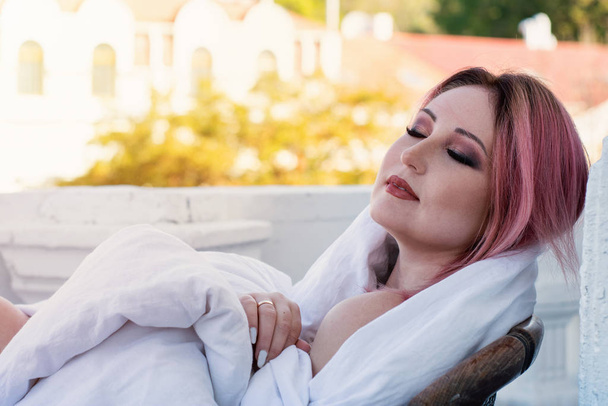 portrait of a beautiful woman with red hair in a white blanket on the balcony on a background of the old town and yellow trees. romantic woman - Photo, Image