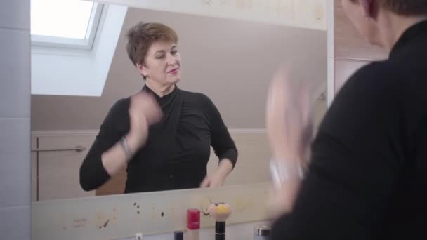 Reflection of happy Caucasian mature woman adjusting her elegant outfit and hair in front of big mirror. Cheerful senior good-looking lady satisfied with her appearance. Fashion, aging, happiness. - Séquence, vidéo