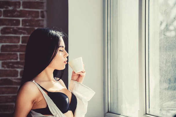 Morning ritual. Girl sexy breasts drinking coffee near windowsill. Attractive female lingerie relaxing at home. Perfect coffee time. But first coffee. Tranquility and peace. Sip of creamy cappuccino - 写真・画像