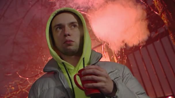 赤いマグカップから熱いお茶やコーヒーを飲む若い男は、夜の風景の背景にタバコを吸う。閉じる-顔、街路灯、霧、バーと壁のアップ. - 映像、動画