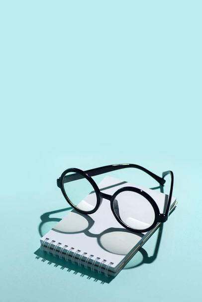 black round glasses lie on a Notepad and cast shadow on the mint background - Zdjęcie, obraz