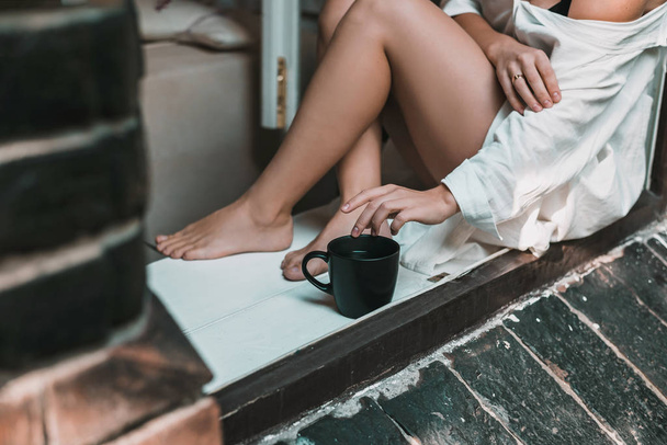 Woman in white dress is sitting on the windowsill with big black tea cup. Morning concept. - Foto, Imagem