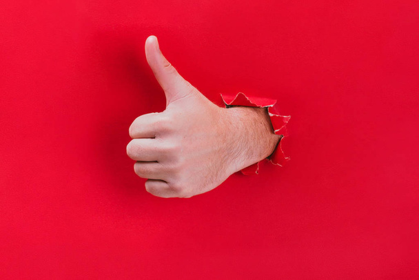 mężczyzna ręka przebija się przez czerwony papier tło i pokazuje jego kciuk w górę. - Zdjęcie, obraz