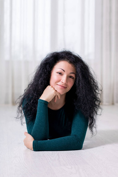 a portrait of a young happy girl with black curly brunette hair who lies on the white floor and holds her head with her hand, in a light interior and dark green pullover. - Photo, Image