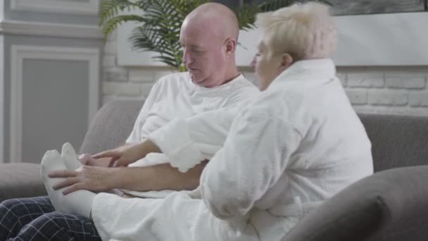 Lähikuva kypsä valkoihoinen mies istuu sohvalla ja hierovat vaimot jalat. Aviomies huolehtii ihastuttavasta puolisostaan. Onnellinen vanhempi pari lepää kotona. Ikuinen rakkaus, hoito, yhdessäolo
. - Materiaali, video