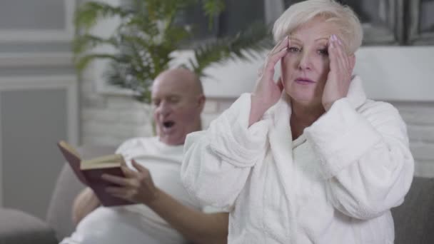 Portrait of sad Caucasian woman with headache sitting in white bathrobe and rubbing temples. Her husband sitting at the background and reading book. Aging, healthcare, eternal love, care. - Metraje, vídeo