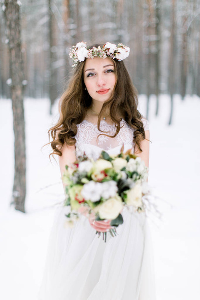 Beautiful bride. Winter wedding. Young woman in wedding dress with a floral cotton head wreath and bouquet standing in the winter forest and looking at the camera - Фото, зображення