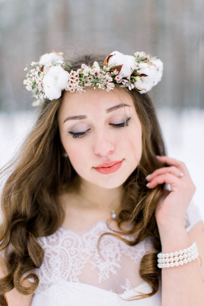 Beautiful bride touching her curly hair, with floral head band wreath, made from flowers and cotton, standing outdoors, on the background of winter forest - Foto, afbeelding