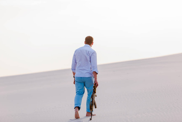man pensive walking walkout bracelet desert shirt blue pans kalashnikov weapon machine-gun sunset white sand.  - Foto, Imagen