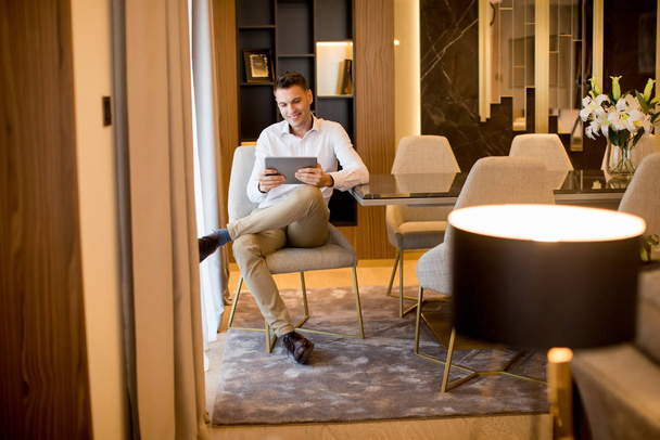 Knappe jongeman zitten en het gebruik van digitale tablet in luxe appartement - Foto, afbeelding