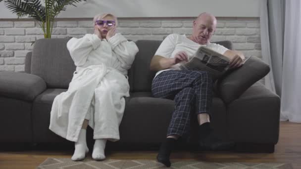 Mature blond Caucasian woman in white bathrobe sitting on sofa and emotionally watching 3D TV. Her baldheaded husband sitting with newspaper and talking to wife. Seniors resting at home. - Imágenes, Vídeo