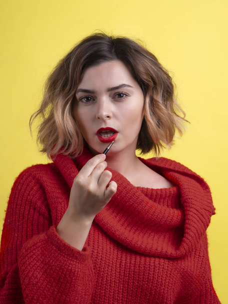 Mujer sexy en rojo aplicando lápiz labial con un cepillo. Cuerpo recortado retrato de primer plano sobre un fondo amarillo. Coqueteando y mirada caliente
 - Foto, imagen