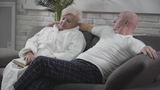 Side view of senior Caucasian bald man sitting on the couch, hugging his wife, talking and gesturing. Mature couple in love spending quiet evening together. Eternal love, togetherness, happiness. - Footage, Video