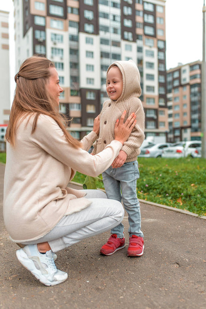 Young happy family, woman mom and little son boy. They relax in autumn on street, smile, walk, and rejoice. Casual warm clothes with a hood jumper. Background building road lawn. - Foto, imagen