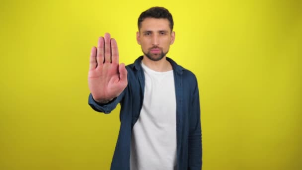 Young man in blue shirt and white T-shirt looks at camera, holds out his hand in front of him and says stop. Stand still. Yellow background with copy space. Stop action. 4k slowmotion footage. - Footage, Video