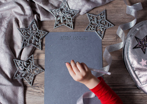 El niño escribe nuevas reglas para el nuevo año en el papel gris. Nuevo plan, palabras, mensaje. Motivación, horario, tarea, nueva vida. Navidad, vacaciones. El fondo plateado. Copos de nieve y juguete de Navidad
 - Foto, Imagen