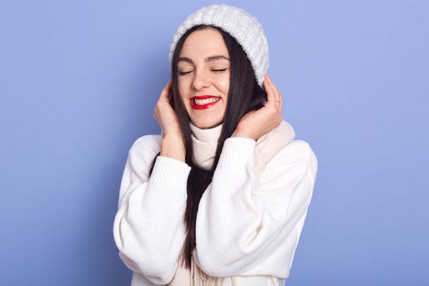 Soñadora guapa morena caucásica joven hembra vistiendo suéter blanco cálido, bufanda y gorra, disfrutando del invierno con los ojos cerrados, aislada sobre fondo de estudio azul. Gente positiva concepto de emoción
. - Foto, imagen