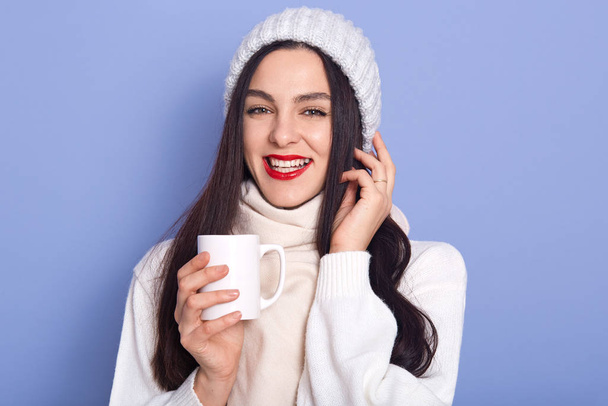 Young attarctive woman holding cap ofhot beverage in her hands, wearing white warm sweater, scarf and cap, posing isolated over blue background, brunette woman with red lips, keeps hand near face - Φωτογραφία, εικόνα