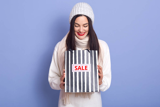 Curious woman with bright red lips and looking in her striped blackand whirte bag with inscription sale. Studio photo of emotional girl wearing white warm clothing isolated over blue background. - Photo, Image