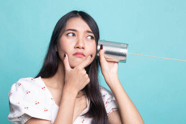Young Asian woman hearing with tin can phone and thinking on cyan background - 写真・画像