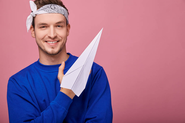 A happy guy in a white with patterns bandana in a dark blue sweater is standing on a pink back with white paper airplane in his hand. Youth, style, confidence, playful mood. Boy smiling. - Photo, image