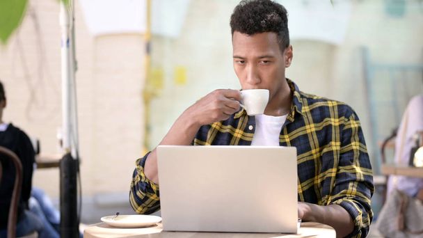 Молодий африканський чоловік п'є каву і працює на ноутбуці, відкритому кафе
 - Фото, зображення