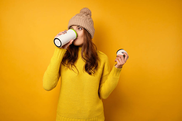 A brunette in a yellow sweater and hat stands on a yellow background and takes a sip from a white thermocup. - Fotoğraf, Görsel